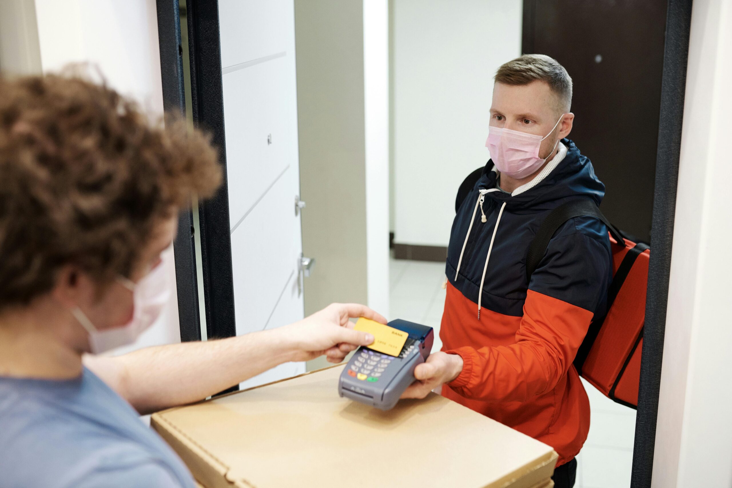Masked deliveryman receiving payment for pizza delivery, highlighting safety measures.
